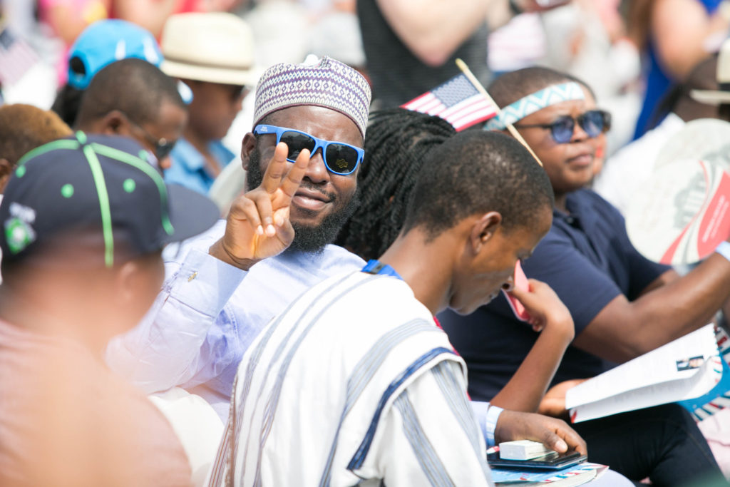 Ahmed, seated in a large outdoor crowd, wearing sunglasses and showing a peace sign with his right hand.